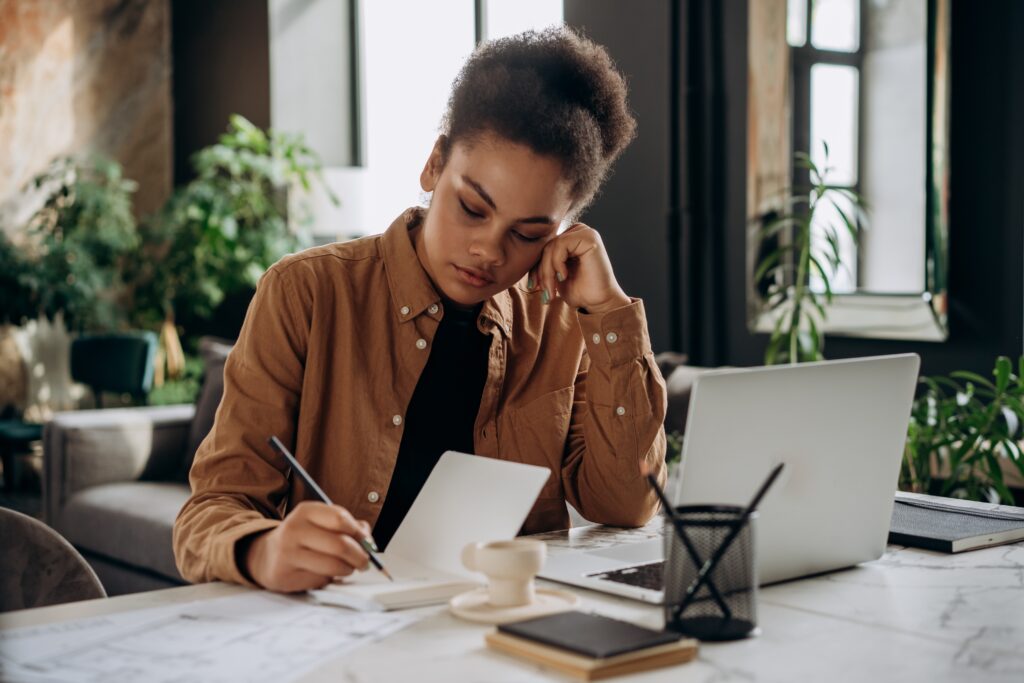 A woman sitting at a desk writing things down on a notepad near her laptop.