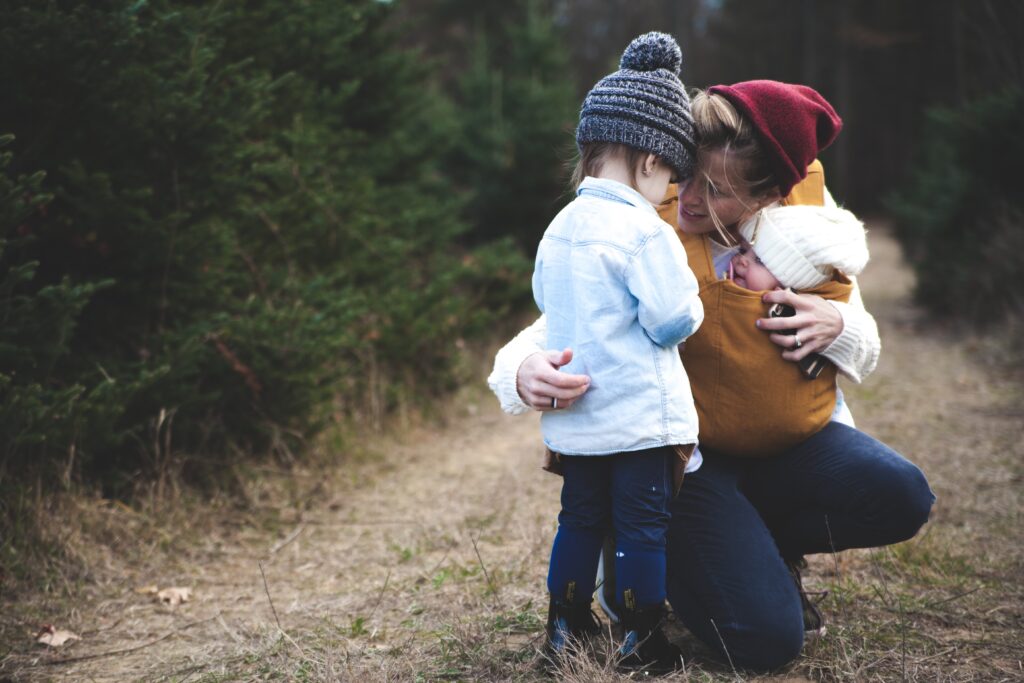 A mother talking to her toddler while her baby is strapped to her chest.