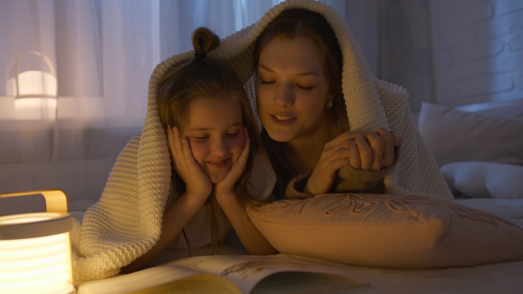 A woman and a little girl covered in a blanket are reading a story.