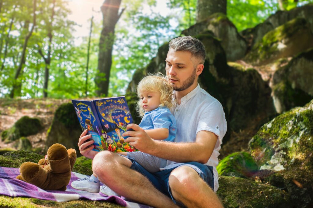 Little girl sitting in her father's lap in the woods and reading a book