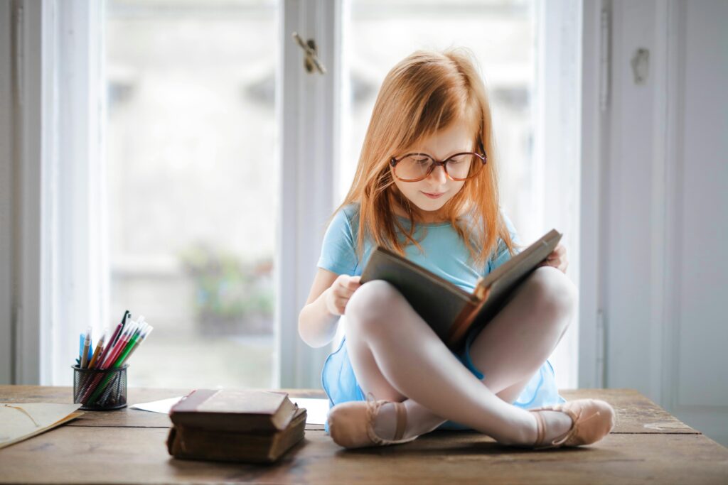 A young girl with glasses reading a book by a window, emphasizing the importance of a reading nook for kids.