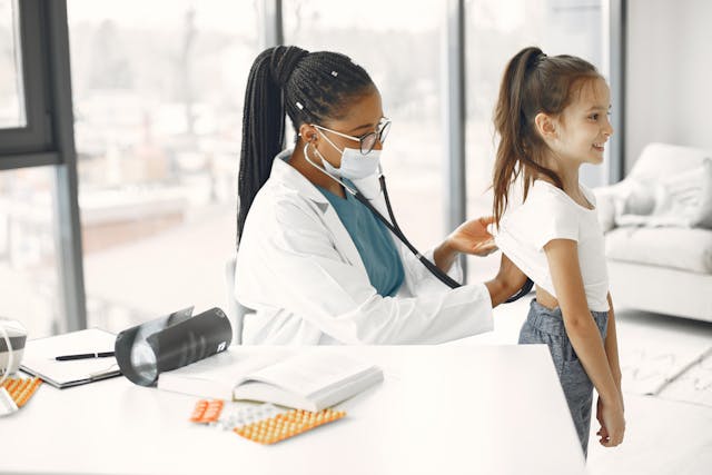 Female doctor using a stethoscope to examine a little girl in a black skirt and a white top.